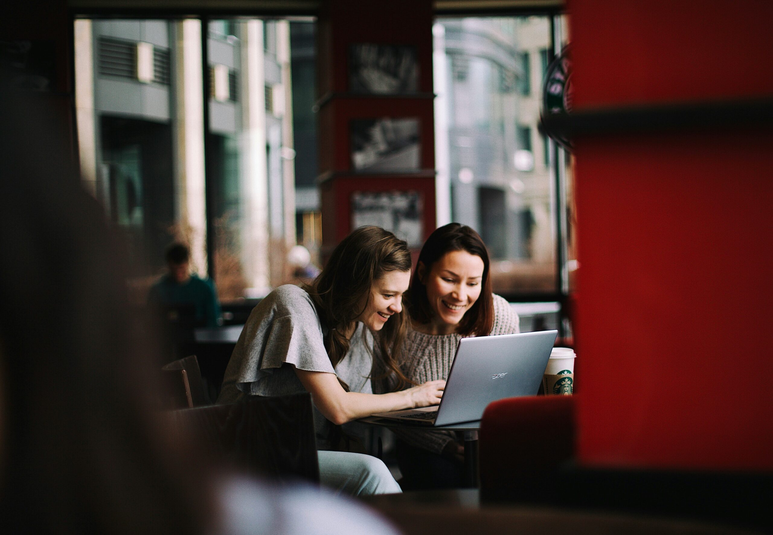 Two women sit in a coworking space looking at a laptop together, smiling.