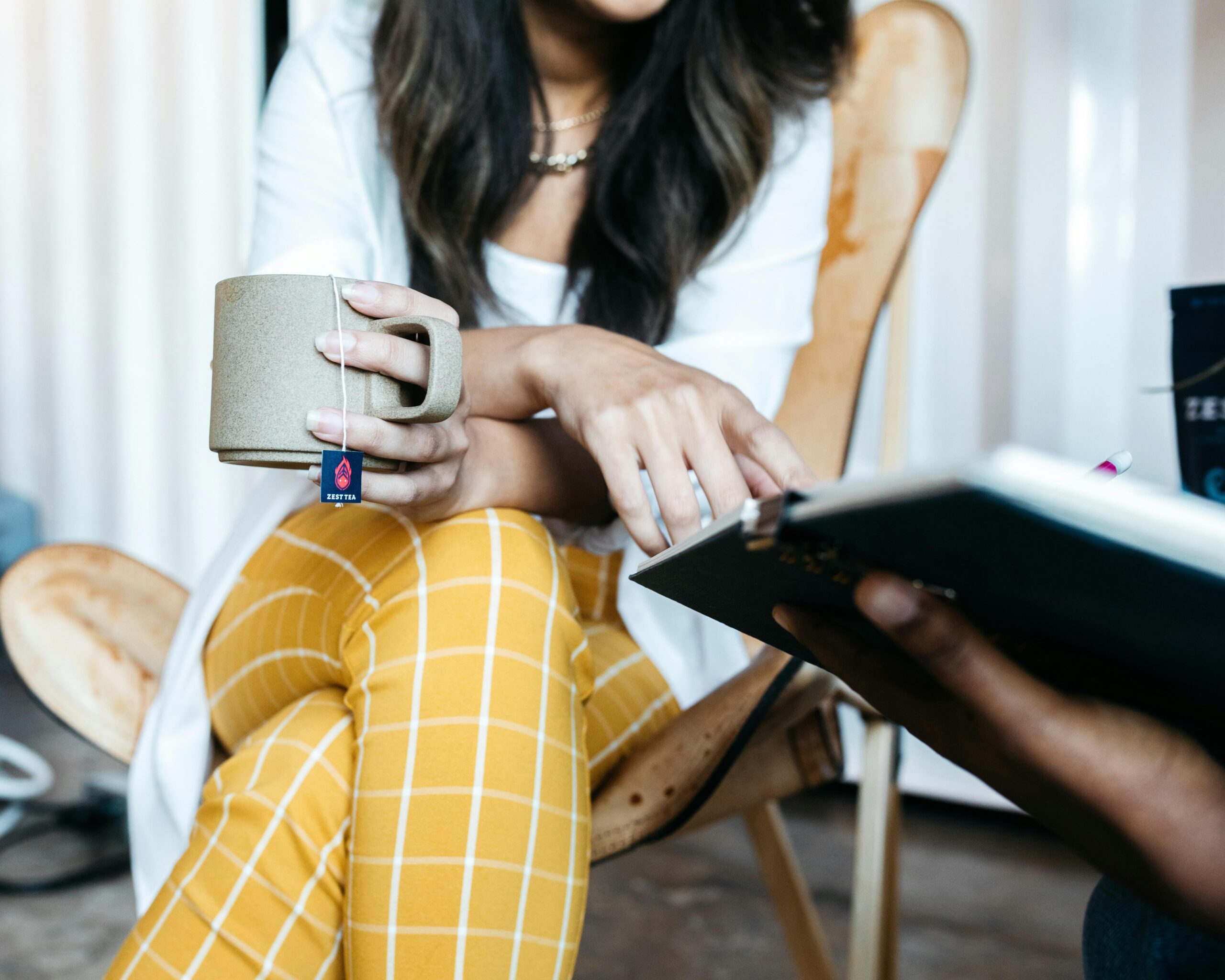 Woman with long dark hair wearing yellow trousers sits in a meeting with a cup of tea