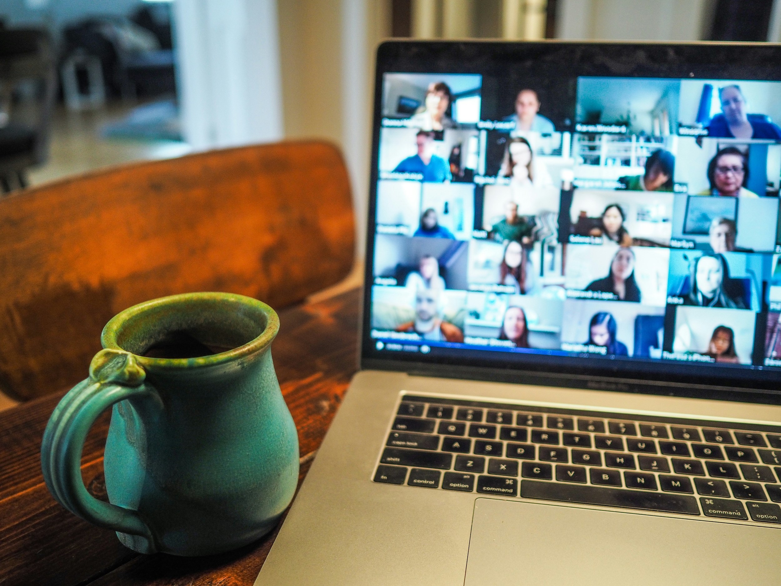 A laptop is open on a table showing multiple meeting attendees online, with a cup of coffee to the left, in a turquoise pottery cup.