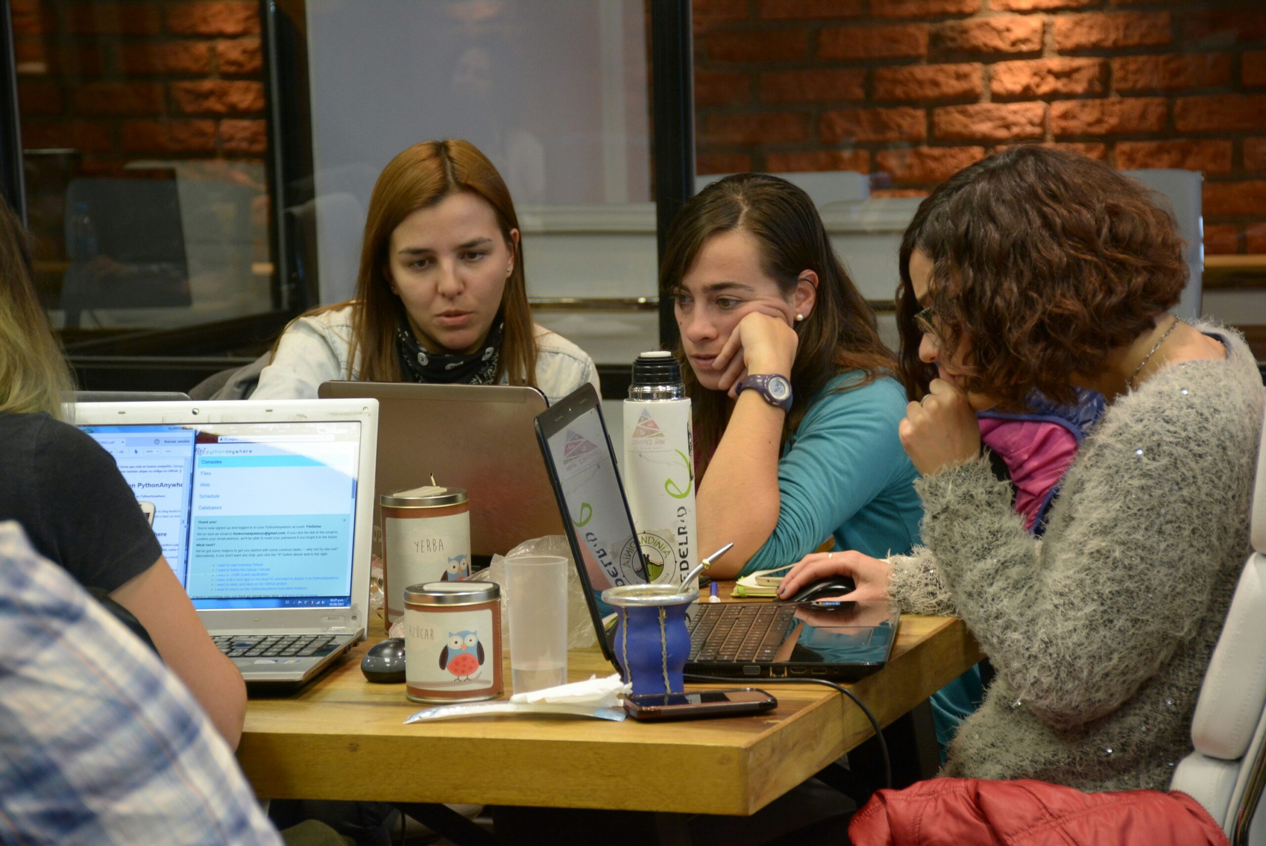 Two young women sit at a desk in a coworking space looking at a laptop together